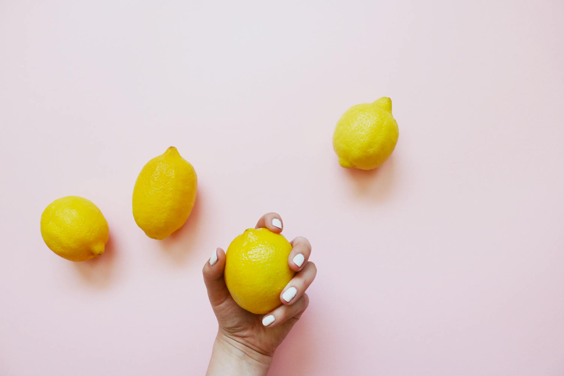 A hand with white nails holding a fresh lemon on a soft pink background, surrounded by three additional lemons.