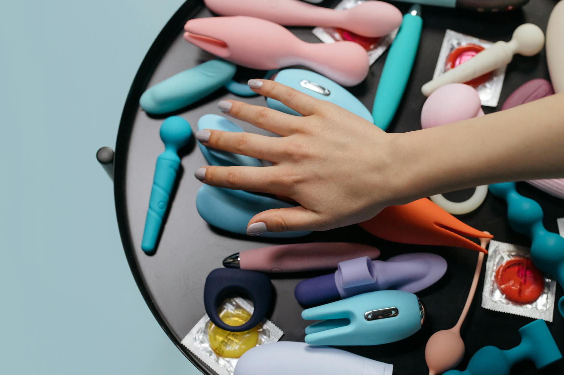 A hand reaching over a variety of colorful clitoral vibrators and sex toys arranged on a surface.