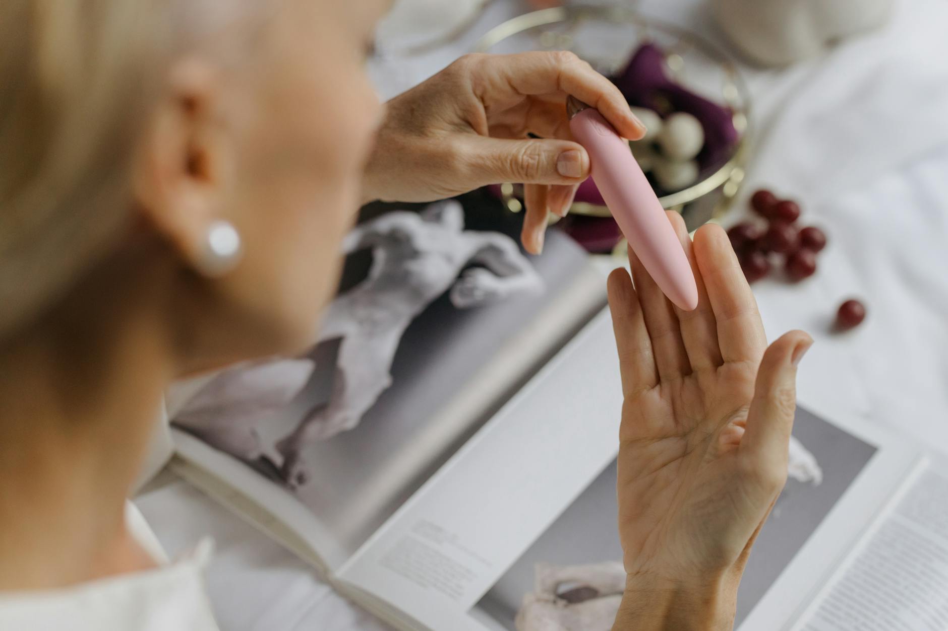 Close-up of a hand holding a pink vibrator with art books in background, representing intimate self-pleasure and rediscovery.